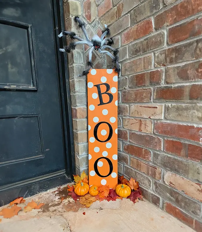 Un letrero de madera naranja con la palabra "BOO" en blanco y negro, decorado con lunares blancos y un gran araña negra. Perfecto para decorar tu puerta o tu jardín en Halloween.
