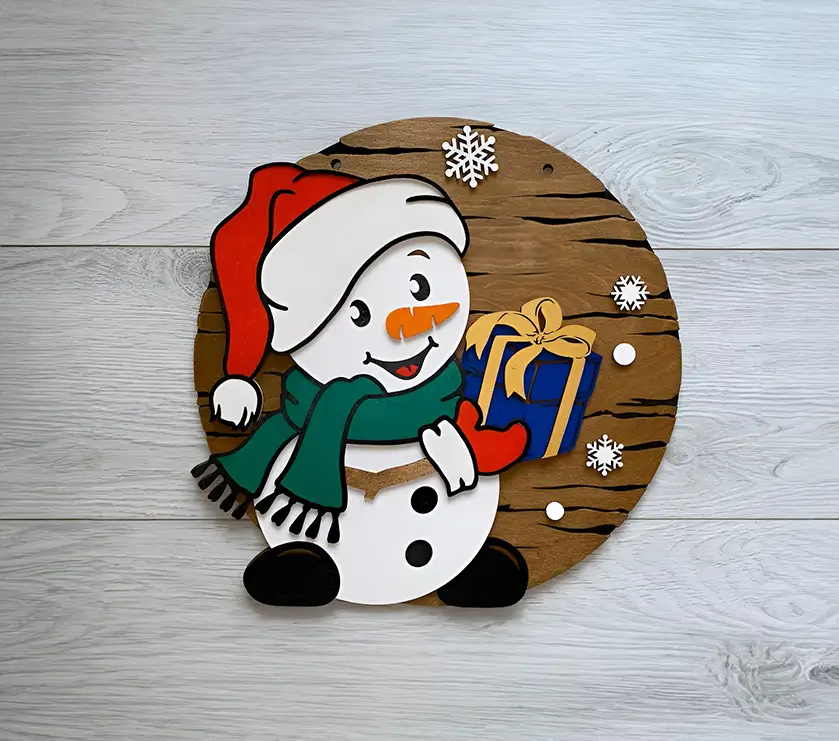 Muñeco de nieve navideño de madera: adorno circular con un diseño clásico y encantador. El muñeco lleva un sombrero y una bufanda, y sostiene un regalo envuelto. Perfecto para regalar o para decorar tu hogar.