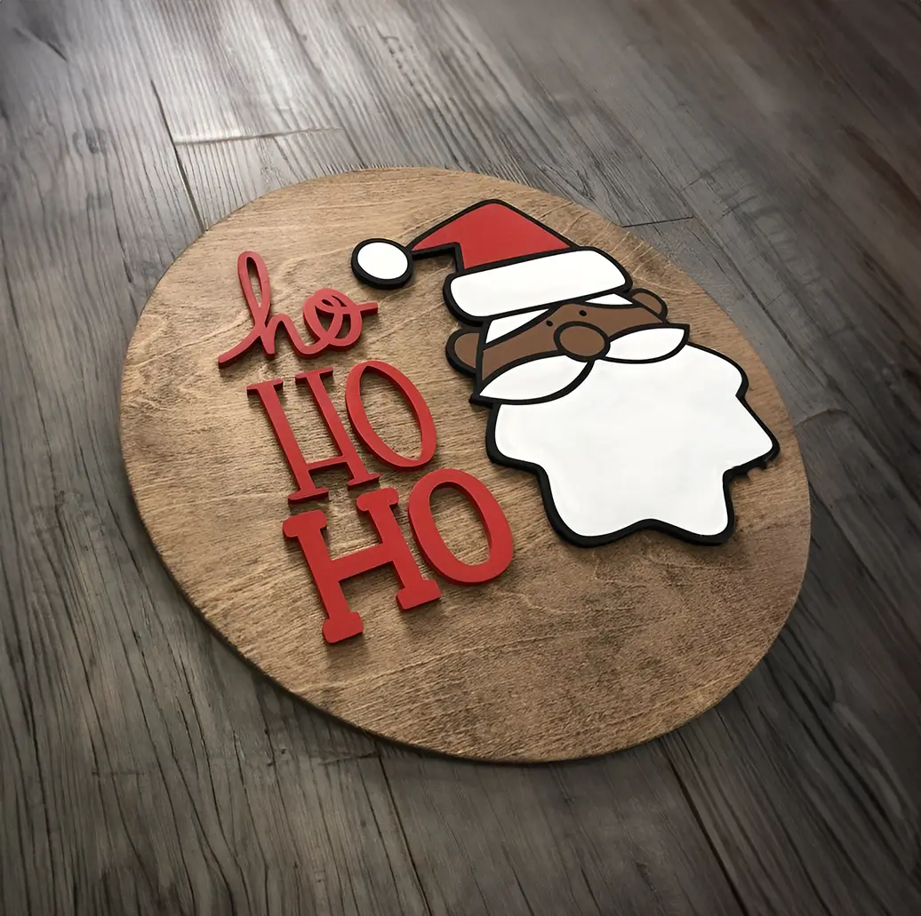 Adorno navideño de madera en forma de círculo con la cara de Santa Claus de piel oscura. El fondo es de madera natural y el texto "Ho Ho Ho" está escrito en rojo.