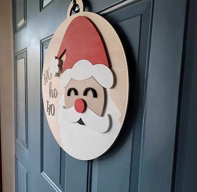 Adorno navideño de madera en forma de círculo con la cara sonriente de Santa Claus. El fondo es de madera natural y el texto "Ho Ho Ho" está escrito en color rojo. Ideal para decorar puertas o paredes durante la temporada navideña.