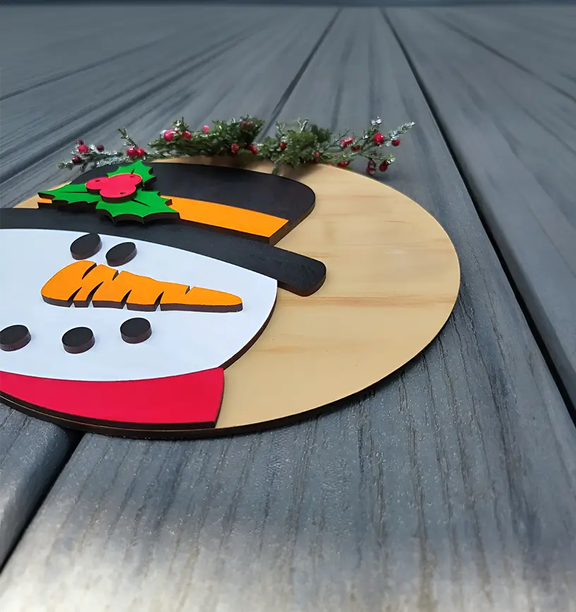 Adorno navideño de madera en forma de círculo con la cara de un muñeco de nieve. El muñeco lleva un sombrero negro, una zanahoria como nariz y botones negros.