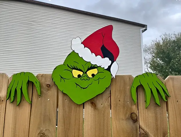 Figura del Grinch artesanal, con expresión traviesa y gorro de Santa Claus, espiando desde una valla de madera en un entorno exterior decorado para las fiestas.