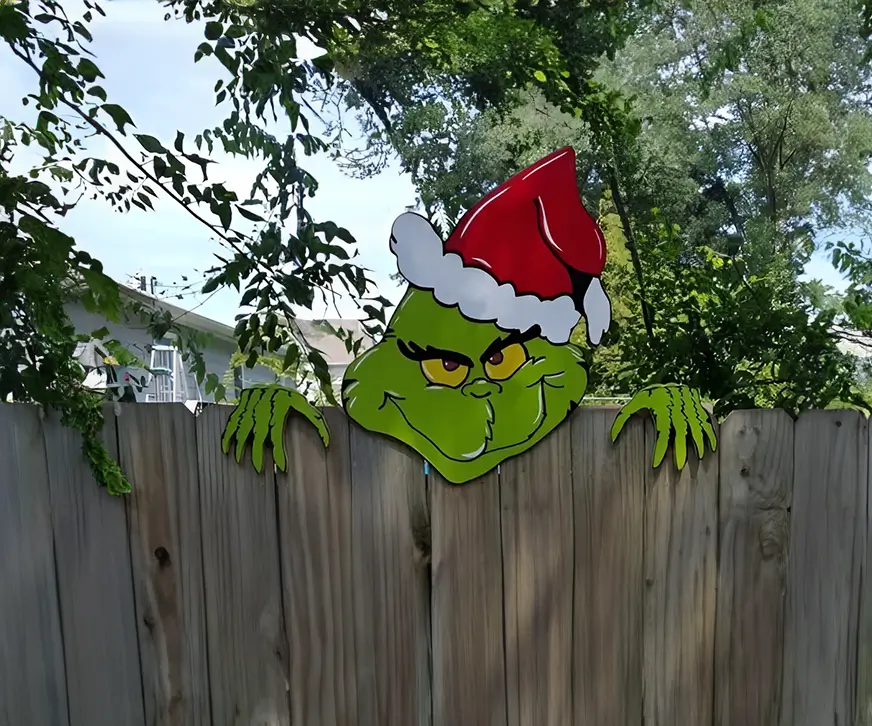Decoración navideña del Grinch hecha a mano, destacando por su diseño colorido y detallado mientras sobresale por encima de una cerca, ideal para jardines o patios.