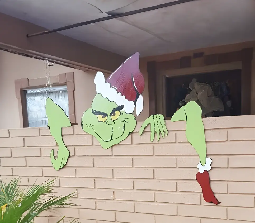 Figura del Grinch decorativa hecha en madera, mostrando su característica expresión traviesa y asomándose desde una barda de ladrillos beige.
