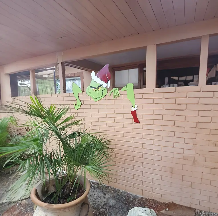 Representación del Grinch con gorro de Santa Claus, sonriendo maliciosamente mientras parece escabullirse detrás de una pared de ladrillo claro.