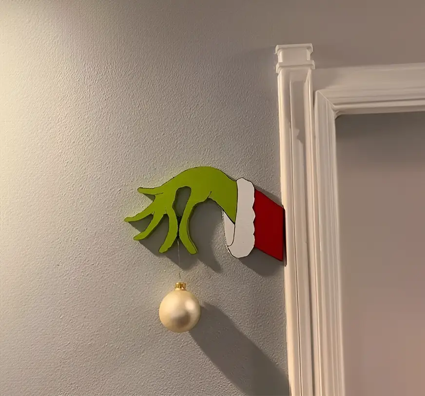 igura decorativa de la mano del Grinch extendida, elaborada en madera cortada a láser y pintada con colores vibrantes. Colocada sobre un piso con acabado de madera clara para una presentación temática navideña.