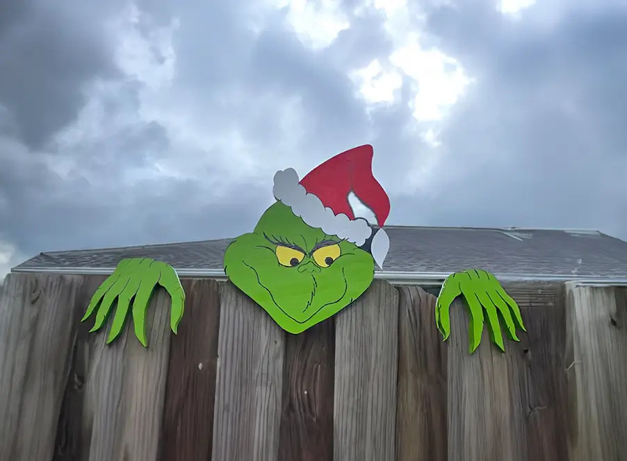 Adorno del Grinch en madera, luciendo un sombrero rojo con detalles blancos mientras se asoma detrás de una cerca de madera natural.