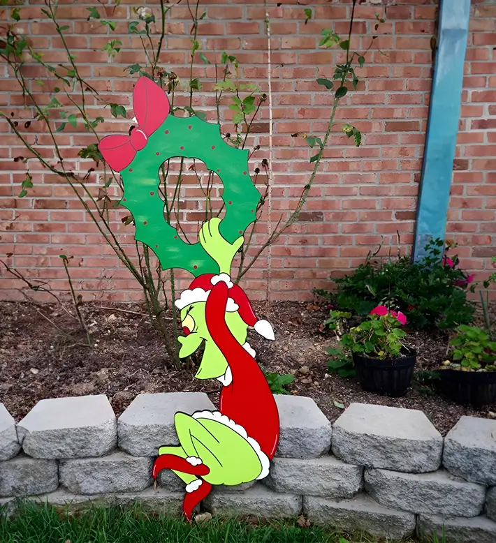 El Grinch, intenta robar una corona navideña roja con un gran lazo. La escena se desarrolla al aire libre, con un fondo de ladrillos y plantas.