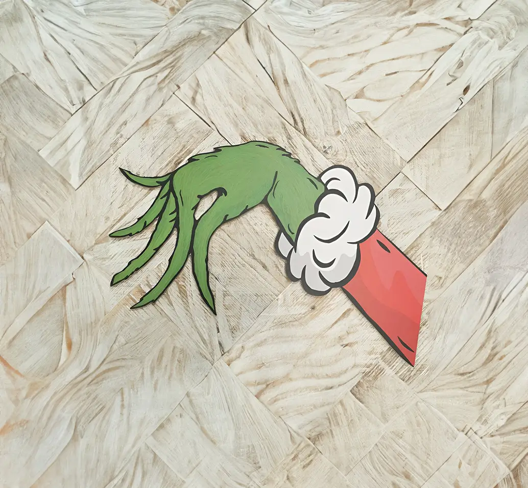 Adorno navideño en madera representando la mano del Grinch en pose característica, con colores detallados y acabados precisos sobre un piso laminado beige.
