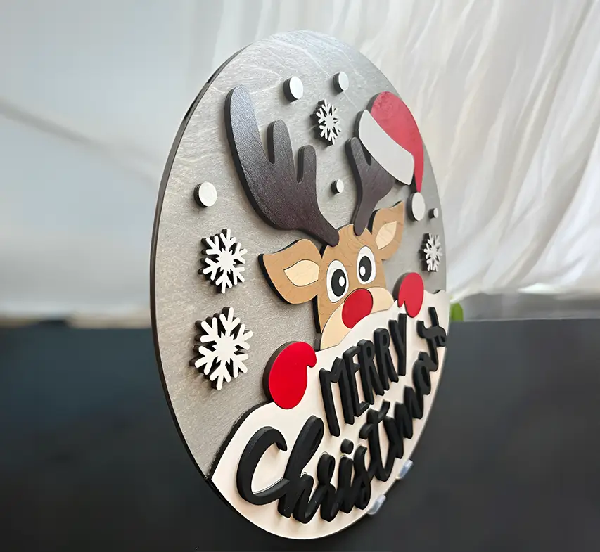Adorno navideño de madera en forma de círculo con la cara de un reno asomando por la parte superior. El reno tiene una nariz roja brillante. En la parte inferior, se puede leer "Merry Christmas" sobre un fondo blanco que simula nieve.